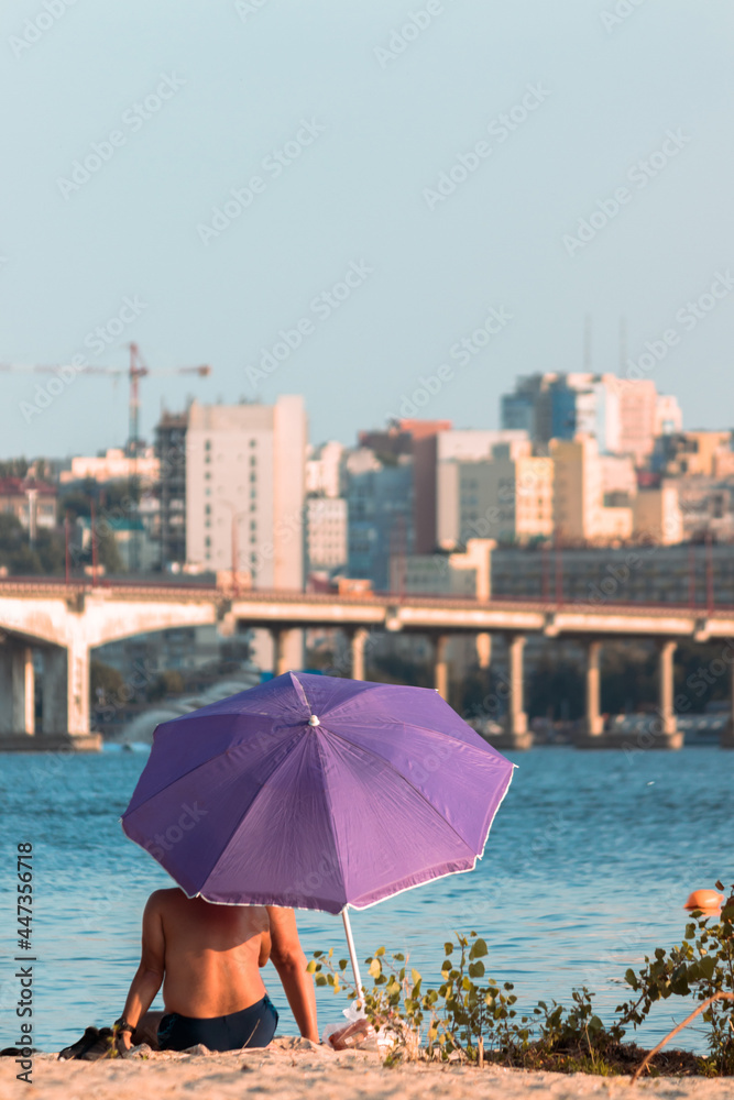 A tanned adult man is resting under a purple umbrella on a city beach with a view of the business center of the city with skyscrapers.