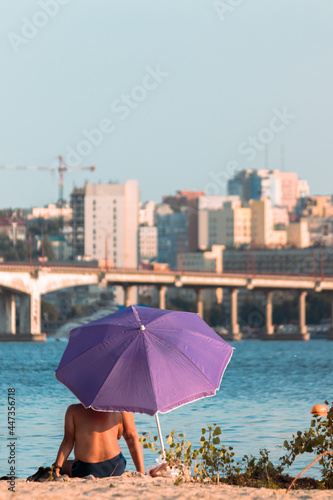 A tanned adult man is resting under a purple umbrella on a city beach with a view of the business center of the city with skyscrapers. © Настя Монастырская