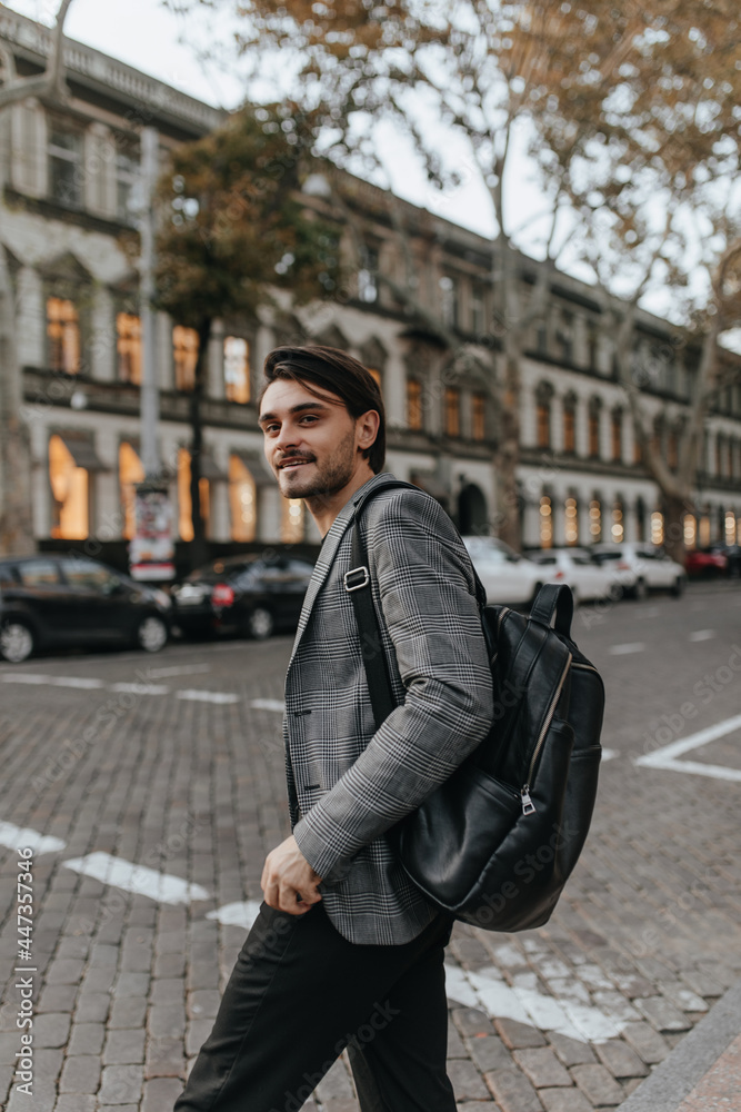 Portrait of gorgeous young gentleman with brunette hair and bristle, grey blazer, dark pants and black backpack, smiling, looking into camera and posing against early autumn street background Portrait of gorgeous young gentleman with brunette hair and bristle, grey blazer, dark pants and black backpack, smiling, looking into camera and posing against early autumn street background