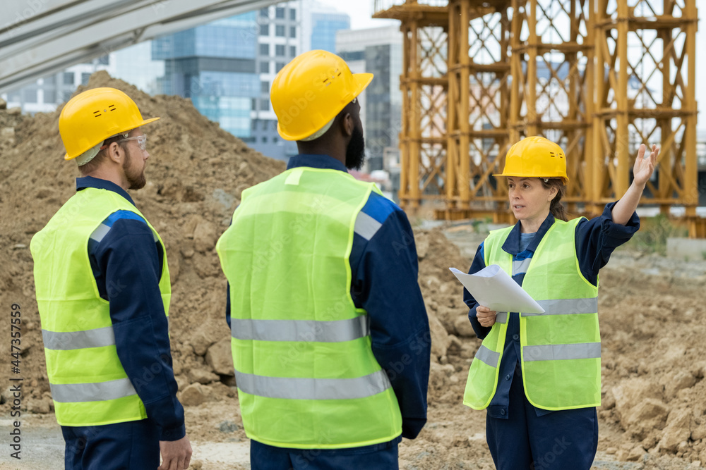 Foreman with blueprint pointing at construction and talking to workers during their work outdoors Foreman with blueprint pointing at construction and talking to workers during their work outdoors