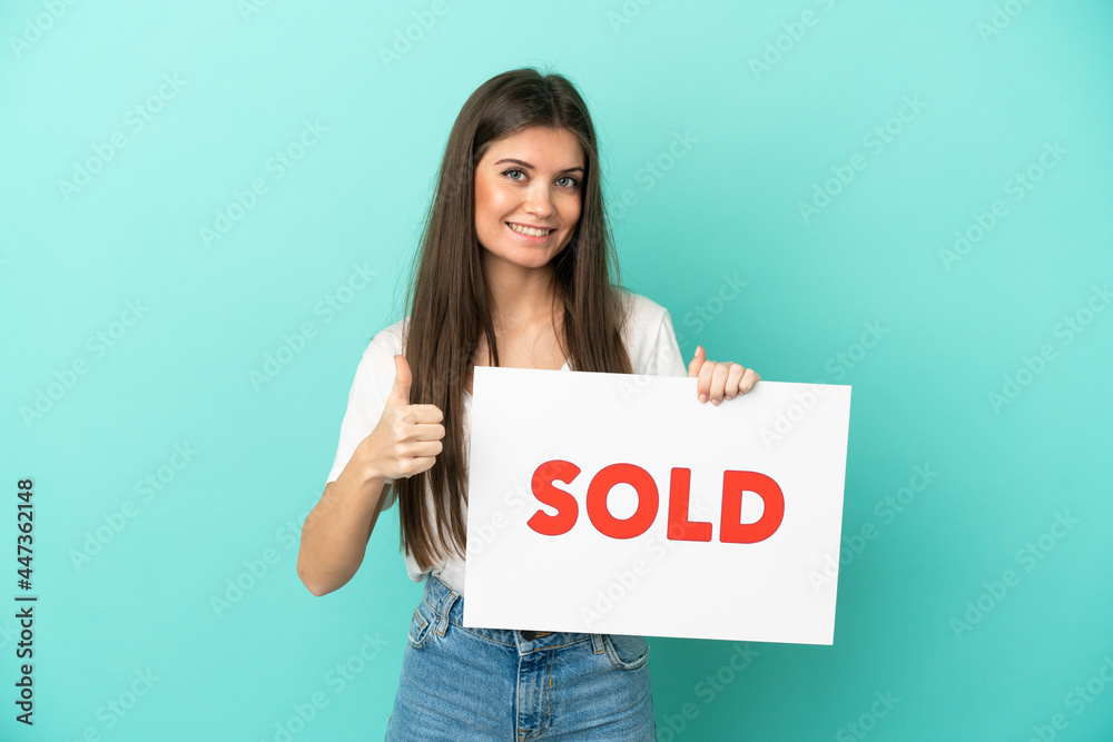 Young caucasian woman isolated on blue background holding a placard with text SOLD with thumb up Young caucasian woman isolated on blue background holding a placard with text SOLD with thumb up