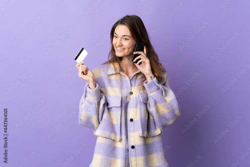 Young Ireland woman isolated on purple background keeping a conversation with the mobile phone and holding a credit card Young Ireland woman isolated on purple background keeping a conversation with the mobile phone and holding a credit card