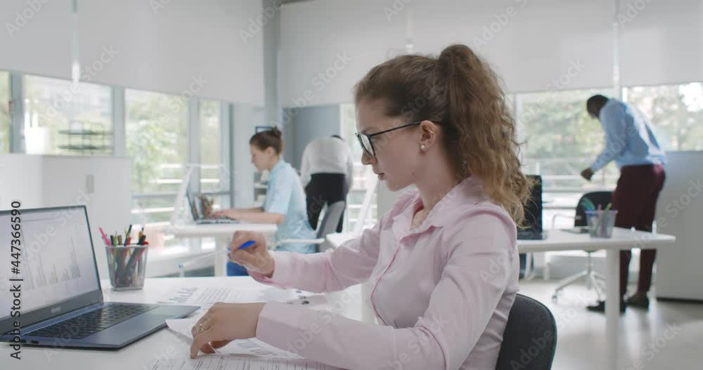 Side view of young businesswoman working on laptop and paperwork in office Side view of young businesswoman working on laptop and paperwork in office