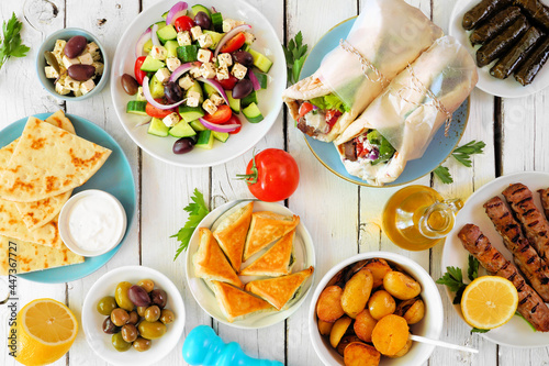 Greek food table scene, above view on a white wood background. Gyros wraps, souvlaki, salad, spanakopita, dolmades, pita and lemon potatoes.