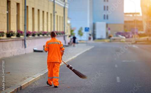 Street cleaner sweeping road and pedestrian zone in evening. Municipal worker sweep road with broomstick and collects garbage. Sanitation worker clean up street. Utility worker cleaning city street