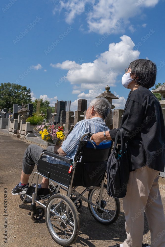Naklejka premium 車椅子に乗った夫を介助しながらお盆の墓参りに向かうマスクをした老夫婦。暑い日差しの照り付ける夏の日に、花を持って墓地を進む（縦）