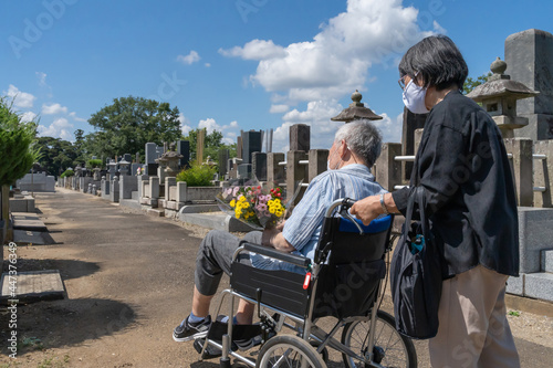 車椅子に乗った夫を介助しながらお盆の墓参りに向かうマスクをした老夫婦。暑い日差しの照り付ける夏の日に、花を持って墓地を進む（横）