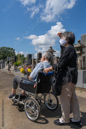 車椅子に乗った夫を介助しながらお盆の墓参りに向かうマスクをした老夫婦。暑い日差しの照り付ける夏の日に、花を持って墓地を進む（縦・日差し）