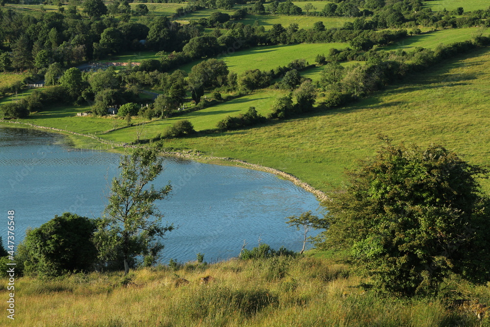 Landscape at Lough Colgagh, County Sligo, Ireland on summer evening featuring lake bordered by hilly farmland fields of pasture in sunlight