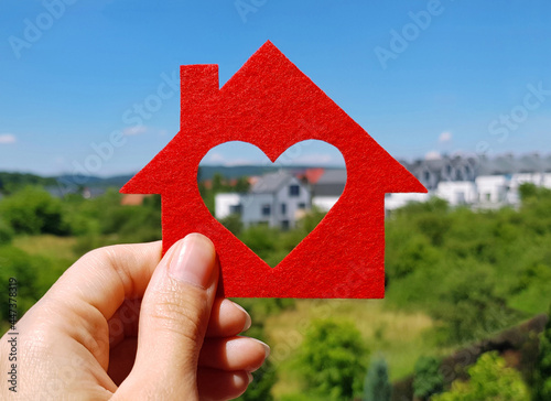 Fototapeta Hand holding red felt house on blurred background of cottage village in summer sunny day