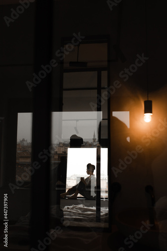 Photo of reflection of woman sitting on windowsill in bedroom. Lady in pajamas and towel on head holds cup.