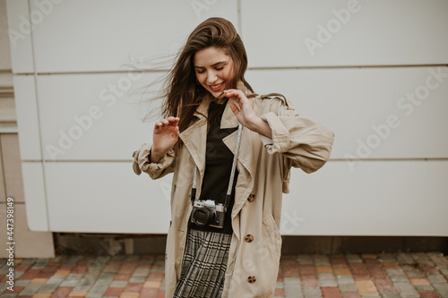 Happy young woman in stylish checkered skirt, black top and trench coat smiles sincerely, looks down and poses outside with retro camera. photo