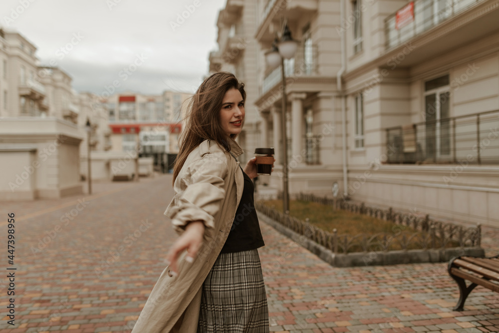 Attractive woman in checkered skirt and long beige trench coat looks into camera, walks outside and holds coffee cup.