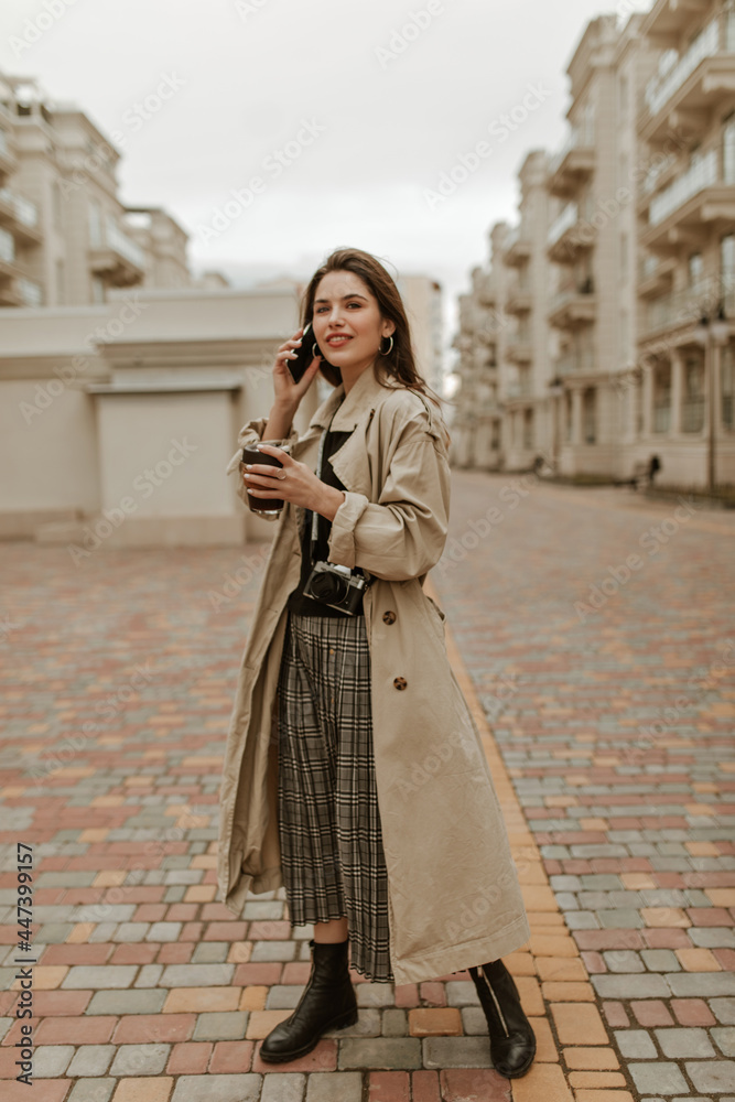 Full-length portrait of brunette charming woman in checkered skirt, black top and beige trench coat talking on phone outside and holding tea cup.