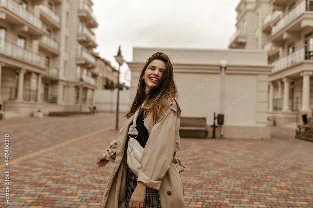 Happy elegant woman in midi beige trench coat smiles sincerely at street. Beautiful brunette lady walks on good mood outside. Happy elegant woman in midi beige trench coat smiles sincerely at street. Beautiful brunette lady walks on good mood outside.