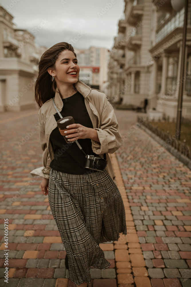Happy brunette woman in checkered skirt, black top and beige trench coat smiles sincerely, holds coffee cup and walks outside. Happy brunette woman in checkered skirt, black top and beige trench coat smiles sincerely, holds coffee cup and walks outside.
