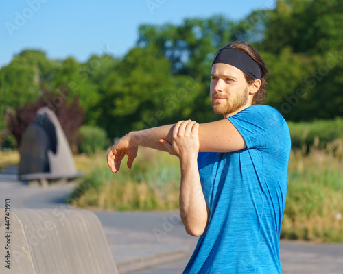 Young handsome man athlete warming up, stretching arm before morning workout in green park, sportive male in blue shirt and headband exercising outdoors on sunny day. Sport and people concept