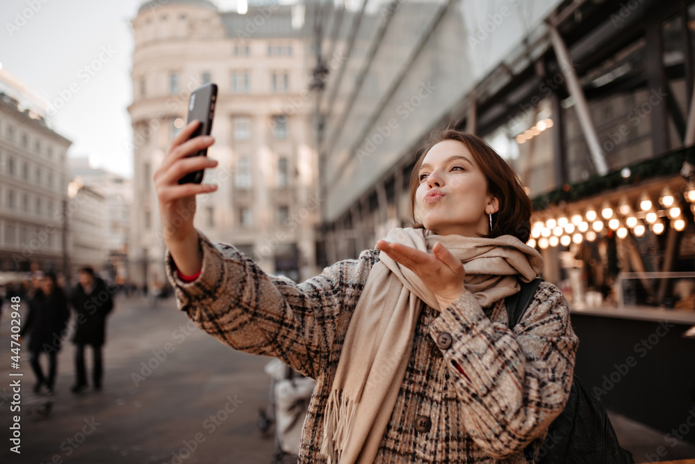 Fototapeta premium Good-humored woman blows kiss and takes selfie outside. Pretty lady in scarf and checkered coat walks outdoors.