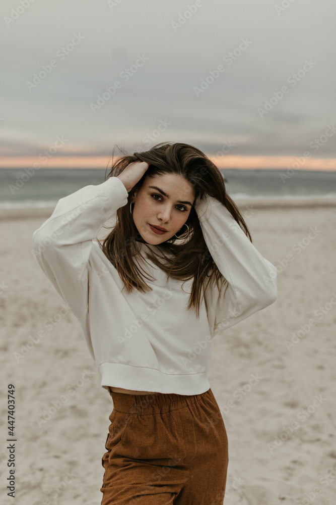 Brunette charming woman ruffles hair and looks into camera. Lady in beige sweater and brown pants poses at beach on sea background. Brunette charming woman ruffles hair and looks into camera. Lady in beige sweater and brown pants poses at beach on sea background.