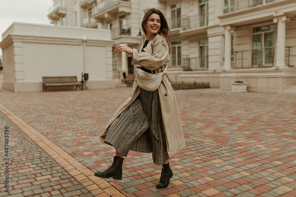 Full length portrait of attractive young woman in beige trench coat walks outside. Pretty lady in midi dress smiles and poses with stylish handbag.