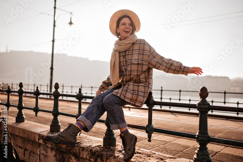 Excited young woman in checkered coat and jeans smiles and sits on fence on quay. Cool girl enjoys sunny autumn day.