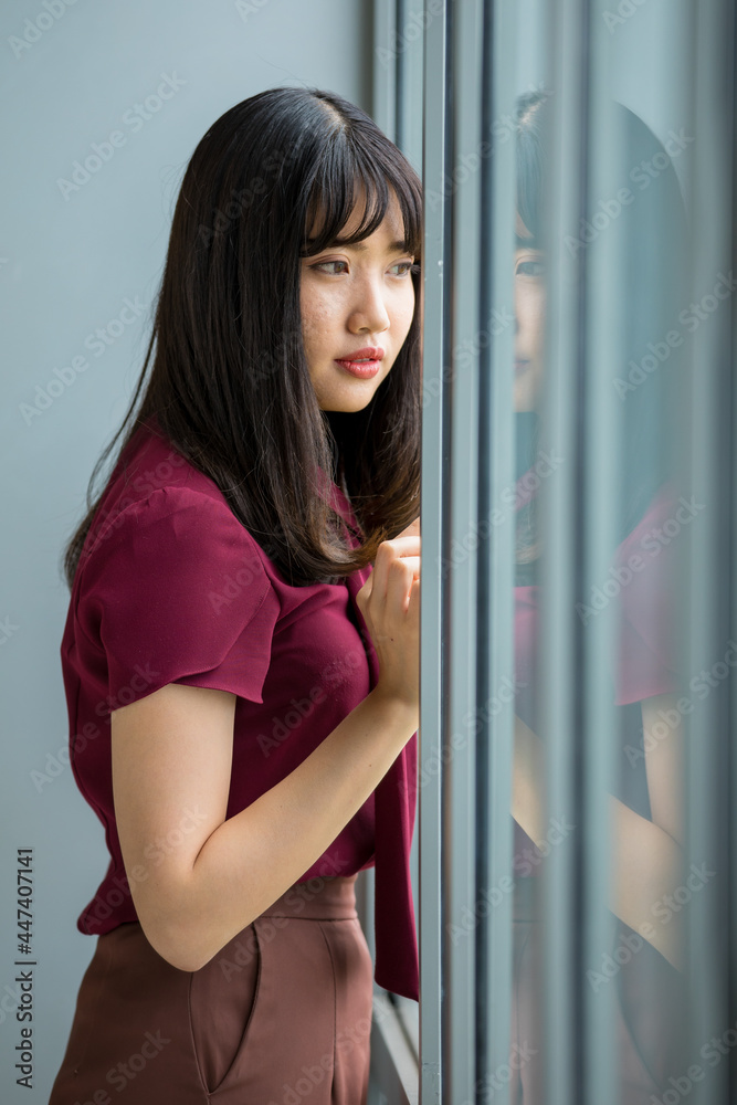 Fototapeta premium Vertical portrait shot of a cute smiling young Asian female in red clothes standing, touching the windows, and looking outside. There is the reflection of woman at the windows