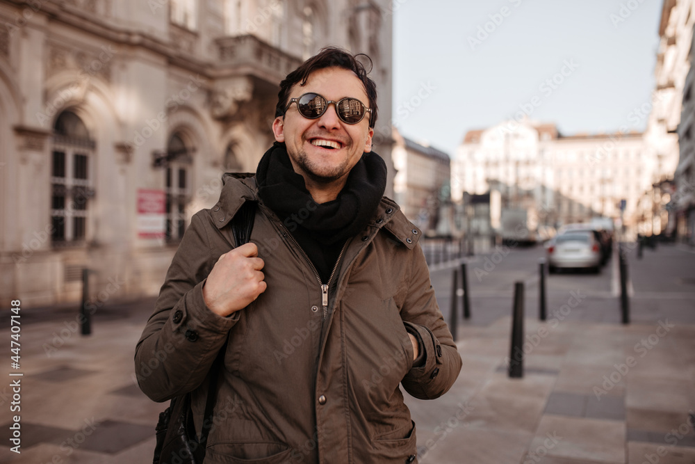 Joyful brunet man in sunglasses, scarf and coat smiles widely outdoors. Good-humored guy walks around city. Joyful brunet man in sunglasses, scarf and coat smiles widely outdoors. Good-humored guy walks around city.