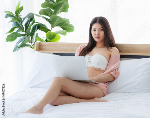 Beautiful young naughty sexy girl on pajamas sit back against pillow on white bed for laptop work and seductively looking out at someone in bright morning of rest day photo