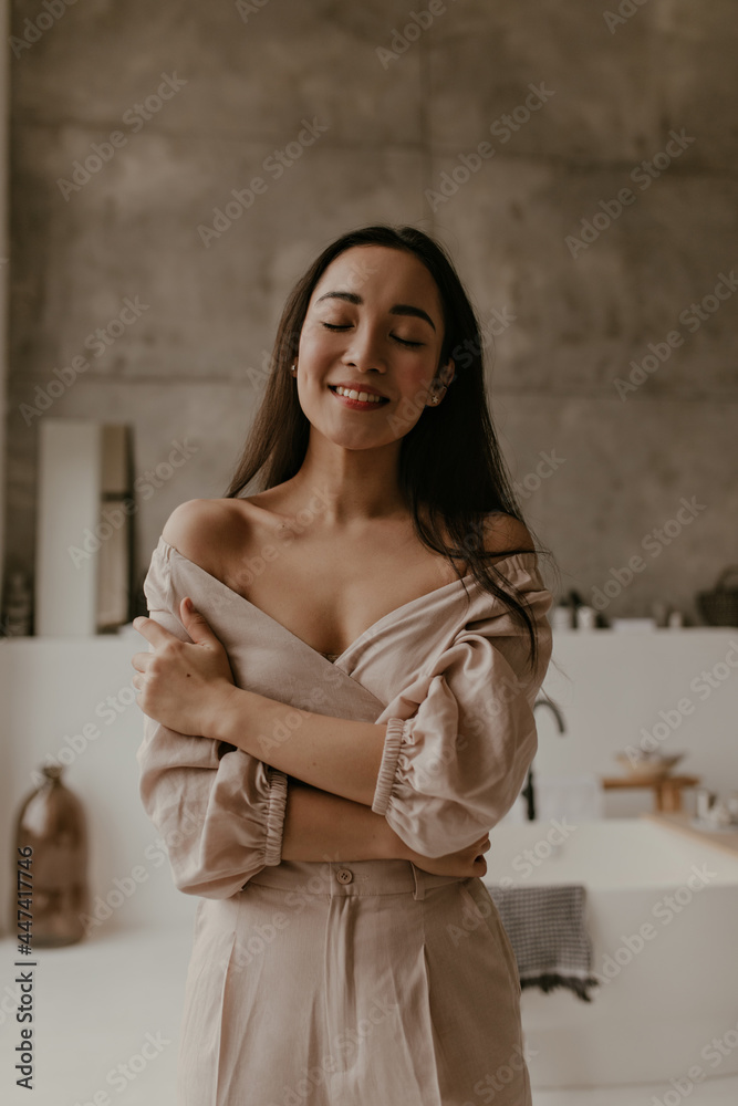 Happy young brunette woman in linen beige outfit smiles sincerely with closed eyes. Asian attractive lady poses in good mood in bathroom.