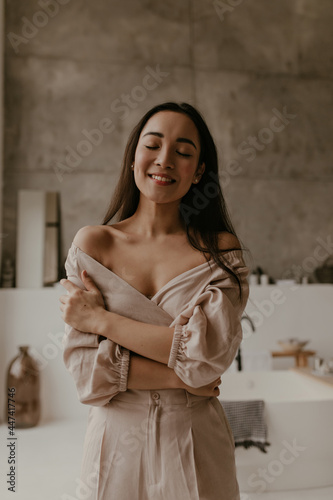 Happy young brunette woman in linen beige outfit smiles sincerely with closed eyes. Asian attractive lady poses in good mood in bathroom.