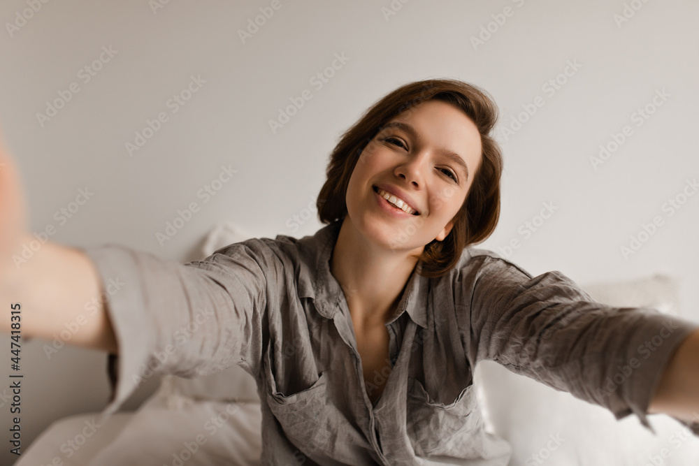 Short-haired woman in grey shirt smiles widely, takes selfie in bedroom. Charming lady in pajamas sits on white bed. Short-haired woman in grey shirt smiles widely, takes selfie in bedroom. Charming lady in pajamas sits on white bed.