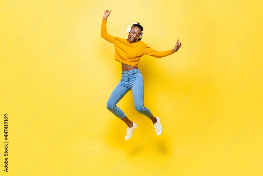 Jumping portrait of happy energetic young African American woman wearing headphones listening to music on yellow isolated studio background
