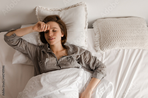 Photo from above of woman sleeping in soft white bed. Charming lady in grey pajama shirt poses in bedroom. © Look!