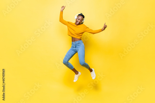 Jumping portrait of happy energetic young African American woman wearing headphones listening to music on yellow isolated studio background photo