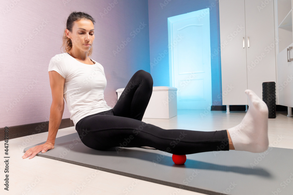Sporty slim caucasian woman doing self massage on fitness mat with massage ball indoors. Self-isolation massage