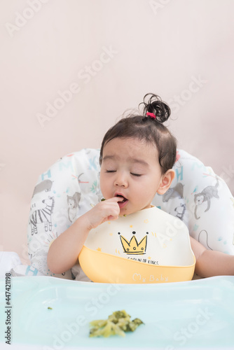 A cute baby girl wearing baby apron sitting on dining table eat broccoli by her self, Baby-Led Weaning concept
