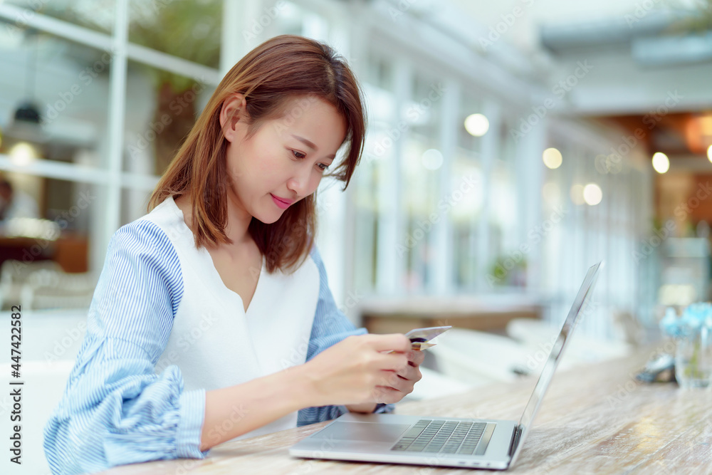 A beautiful Asian woman in a white shirt is sitting in front of a computer with a credit card in her hand to prepare for online shopping. Her face looks very happy in a bakery. A beautiful Asian woman in a white shirt is sitting in front of a computer with a credit card in her hand to prepare for online shopping. Her face looks very happy in a bakery.