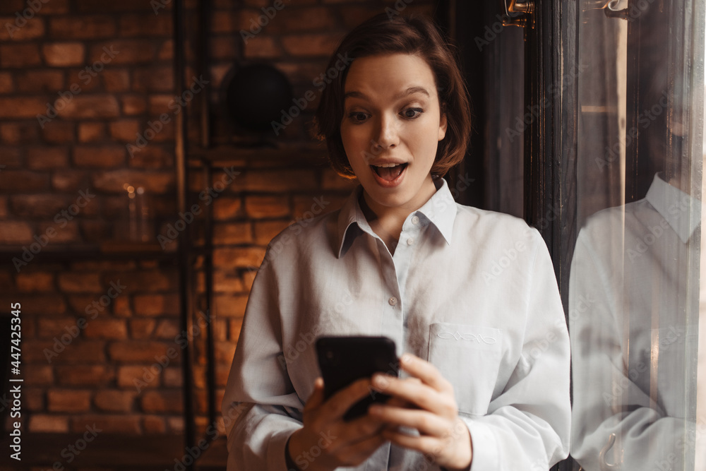Fototapeta premium Excited surprised woman in blue shirt looks at phone. Cheerful lady in blouse stands nearby window in cozy room.