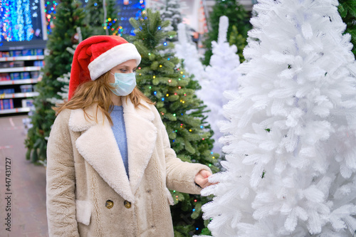 A woman in a medical face mask chooses a faux Christmas tree in a store with New Year gifts photo