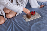 Young man dips strawberries in chocolate paste in a glass jar on a marble tray, feeds a woman. Lovers in bathrobes have breakfast in blue bed in the morning. A loving couple travels honeymoon
