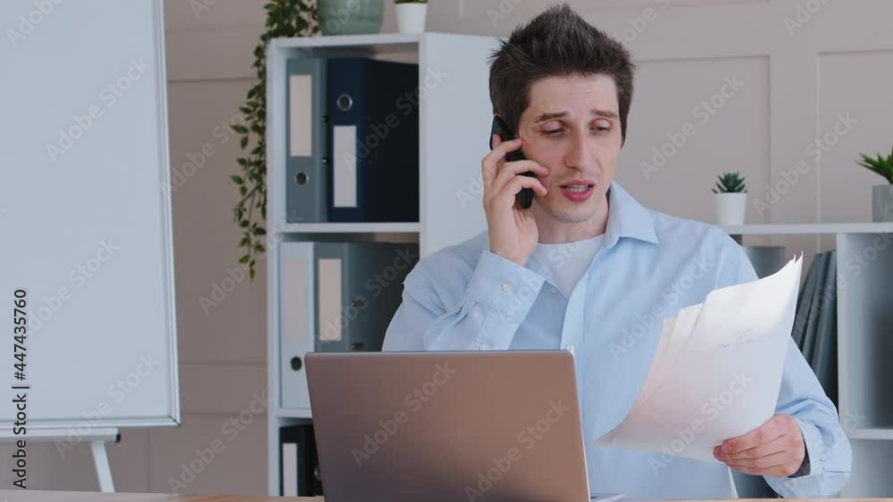 Concentrated emotional business man caucasian male worker manager sitting at office table with laptop holding papers in hands documents talking mobile phone answering call consulting with smartphone Concentrated emotional business man caucasian male worker manager sitting at office table with laptop holding papers in hands documents talking mobile phone answering call consulting with smartphone