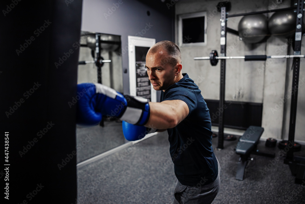 Boxing training in the gym. A man wearing gloves and hitting a black punching bag in a boxing gym with a mirror. A dedicated strong man dressed in sports clothes punching a bag, arm workout Boxing training in the gym. A man wearing gloves and hitting a black punching bag in a boxing gym with a mirror. A dedicated strong man dressed in sports clothes punching a bag, arm workout