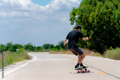 A back view of a young Asian man wearing a black shirt and pants is skateboarding on a rural road., Play surf skate.