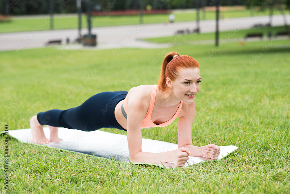 Redhead caucasian girl standing in a plank on a mat on the grass in the park