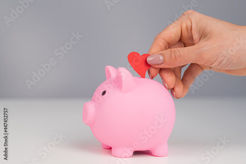 Faceless caucasian woman folds a small heart into a pink piggy bank on a white background. Love saving concept