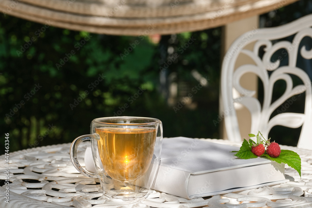 Transparent glass cup of herbal tea against the blurred background of a white garden table in the garden. White book with red raspberries. Shallow depth of field