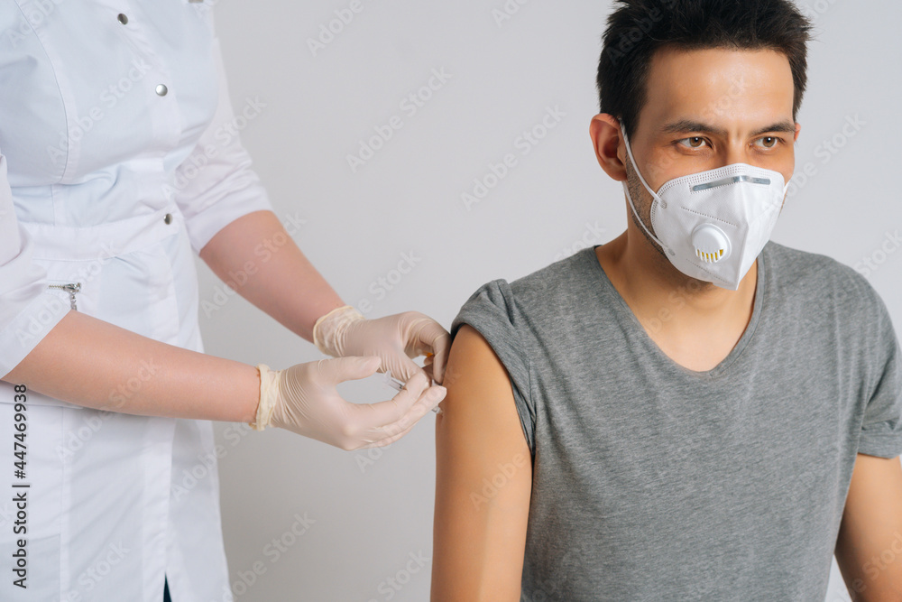 Close-up studio shot of young man wearing face mask receiving vaccine against coronavirus sitting on chair on isolated white background. Portrait of nurse using syringe to inject vaccine to patient.