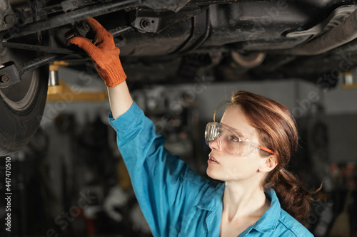 Cavsian woman auto mechanic at a car service station stands under a raised car and inspects the chassis. dressed in a blue overalls.