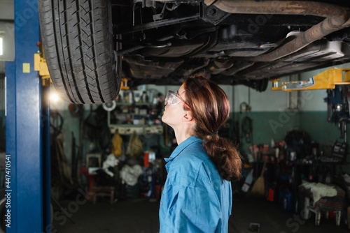 portrait of a professional female auto mechanic working under a vehicle on a lift in service. makes a car inspection. The specialist wears protective goggles.