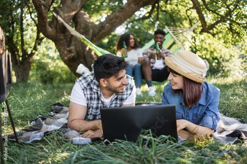 Young indian guy and caucasian woman lying on soft plaid at garden and using wireless laptop. African american couple relaxing in hammock with beer in hands. Four happy friends on picnic.